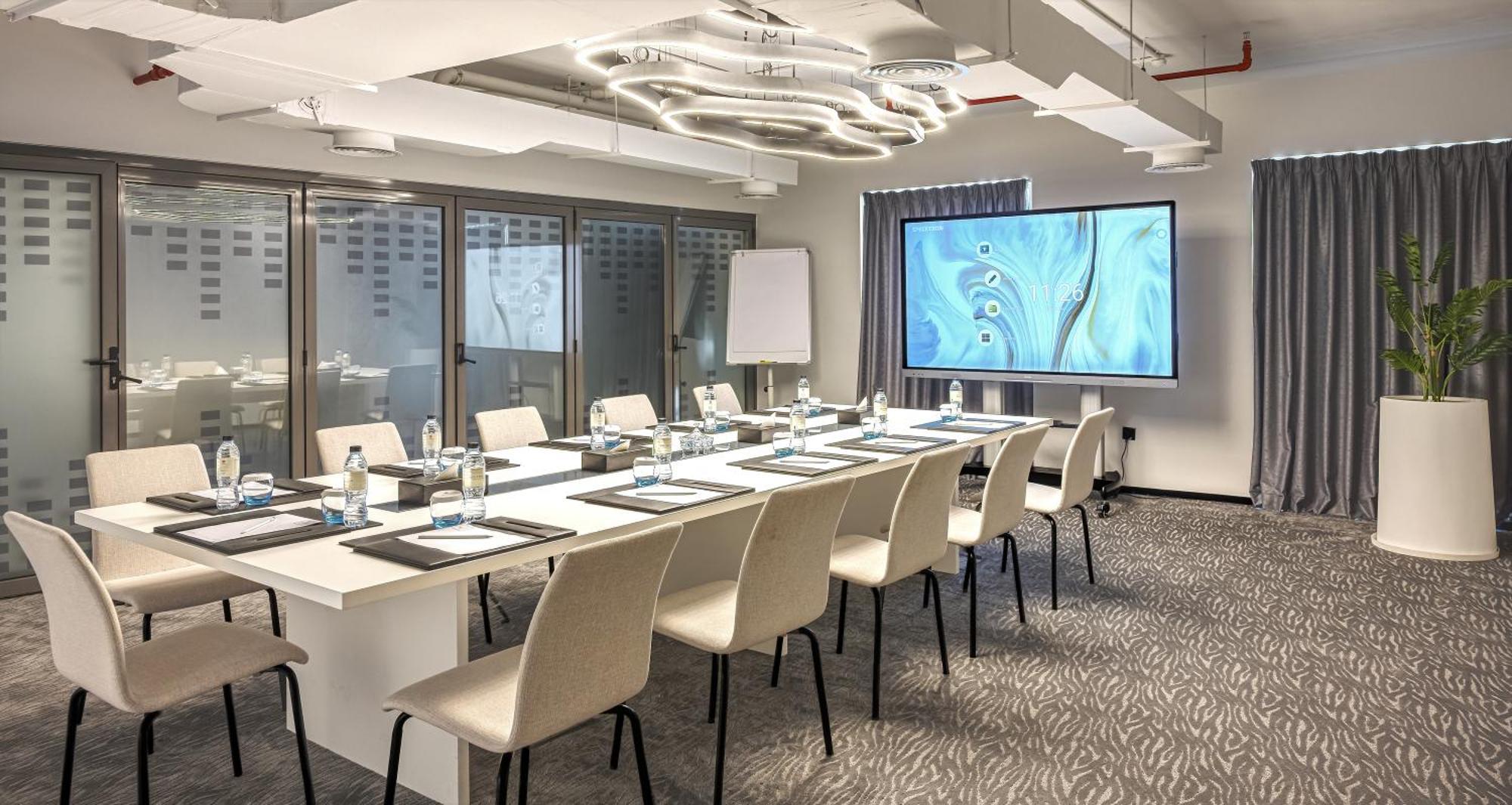 มิลเลนเนียม เอเตรีย บิสซิเนส เบย์ Aparthotel ดูไบ ภายนอก รูปภาพ The photo shows a modern conference room set up for a meeting. There is a long table with several chairs arranged around it, each with a notepad and pen. A large screen displays a graphic or presentation, and there is a whiteboard in the corner. The 