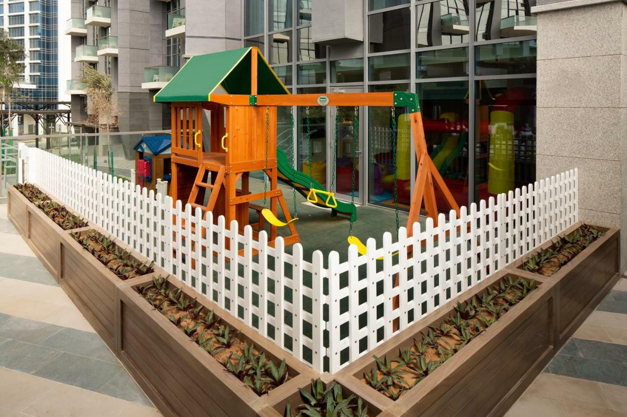 มิลเลนเนียม เอเตรีย บิสซิเนส เบย์ Aparthotel ดูไบ ภายนอก รูปภาพ The image shows a children's playground area, which is enclosed by a white picket fence. The playground features a wooden play structure with a slide, swings, and climbing elements. Surrounding the play area are planters with greenery, and the backdr