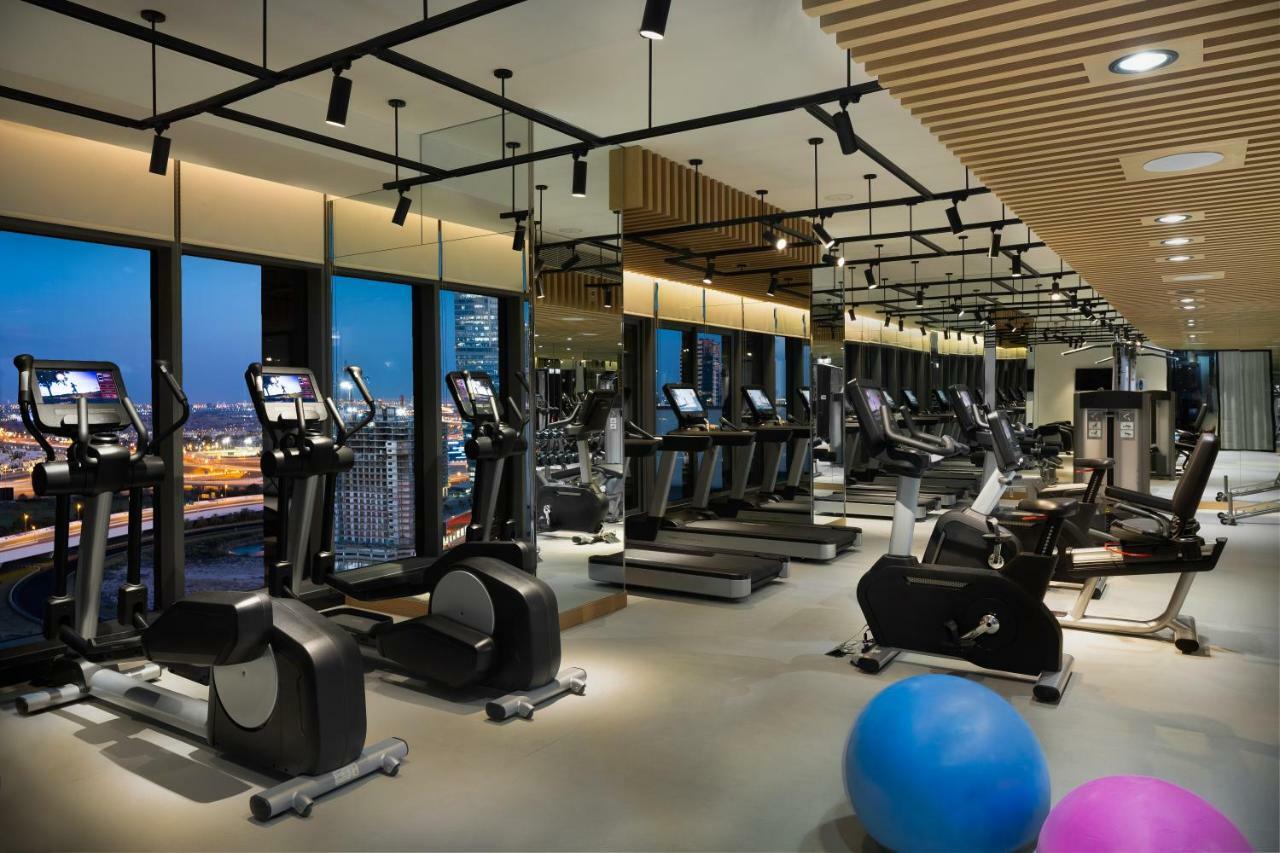 มิลเลนเนียม เอเตรีย บิสซิเนส เบย์ Aparthotel ดูไบ ภายนอก รูปภาพ The photo shows a modern gym with various types of exercise equipment available. There are several treadmills and elliptical machines arranged in rows, with large windows in the background providing a view of the city. The lighting is bright and cont