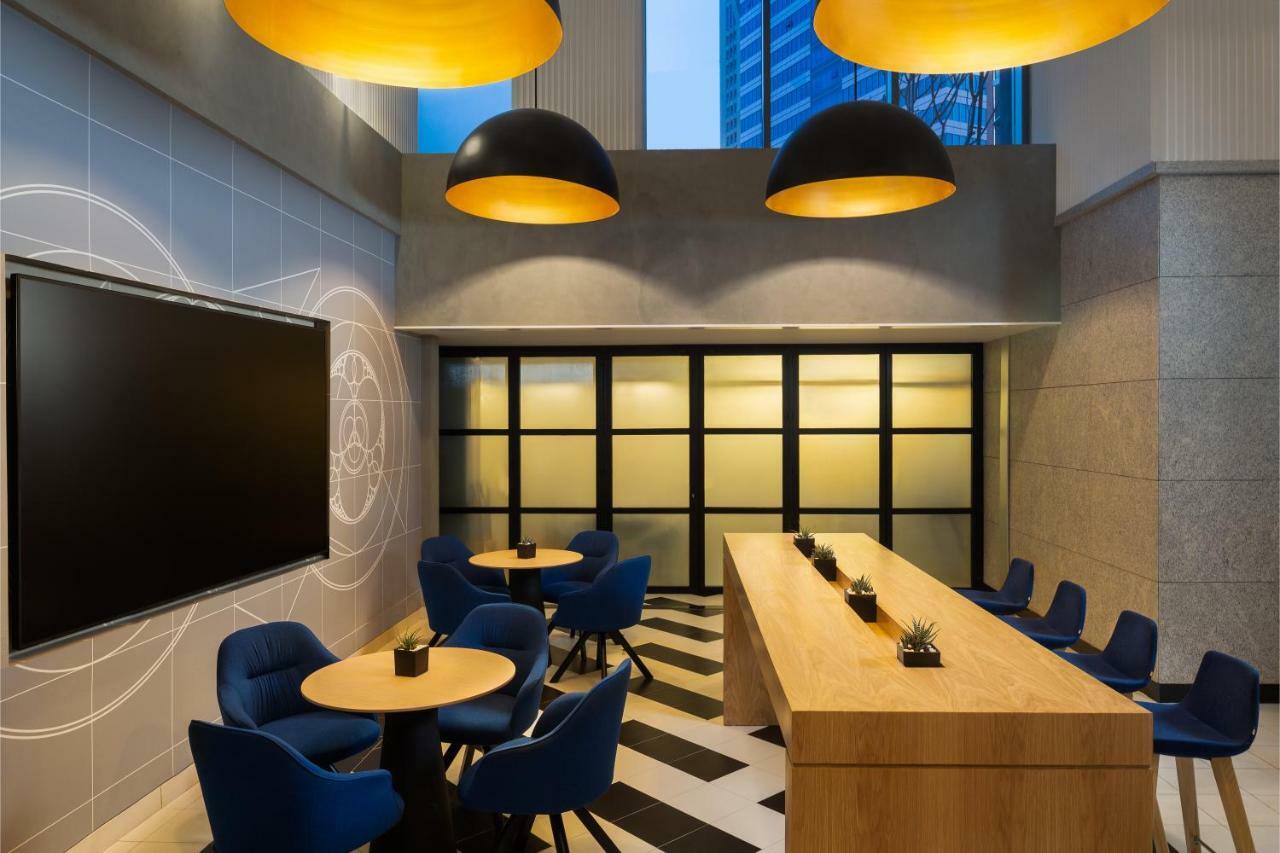 มิลเลนเนียม เอเตรีย บิสซิเนส เบย์ Aparthotel ดูไบ ภายนอก รูปภาพ The photo shows a modern interior space, likely a lounge or meeting area. It features a long wooden table with some small plants on it, accompanied by several blue, circular chairs arranged around it. There is a large flat-screen television mounted o