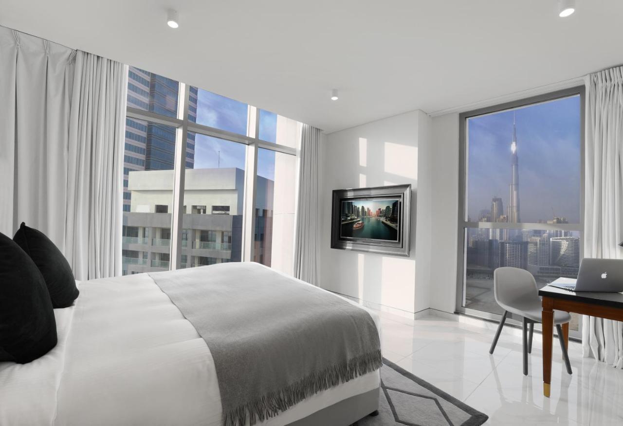 มิลเลนเนียม เอเตรีย บิสซิเนส เบย์ Aparthotel ดูไบ ภายนอก รูปภาพ The photo depicts a modern hotel room that features a large bed with a gray throw blanket. There are large windows allowing natural light to fill the space, showcasing a city skyline view. A flat-screen television is mounted on the wall opposite the 